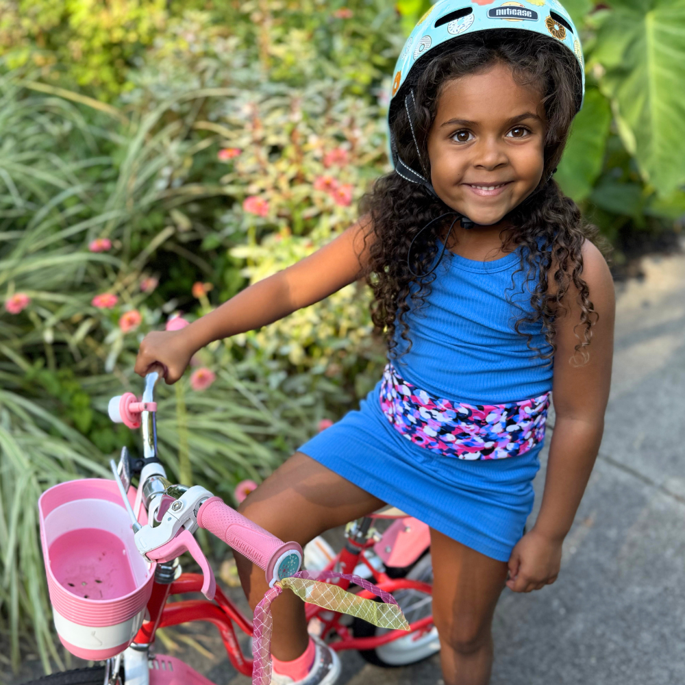 Young girl in a blue dress and helmet standing next to a pink bicycle outdoors and wearing a pink blue and white fanny pack