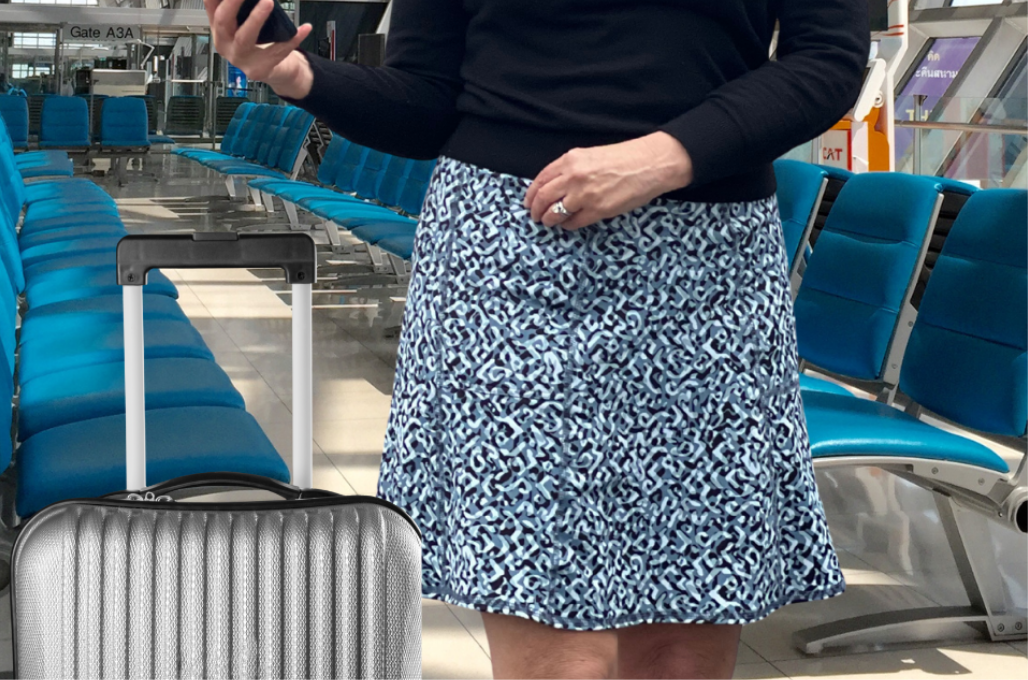 Person in a grey blue patterned skirt standing in an airport with a suitcase.