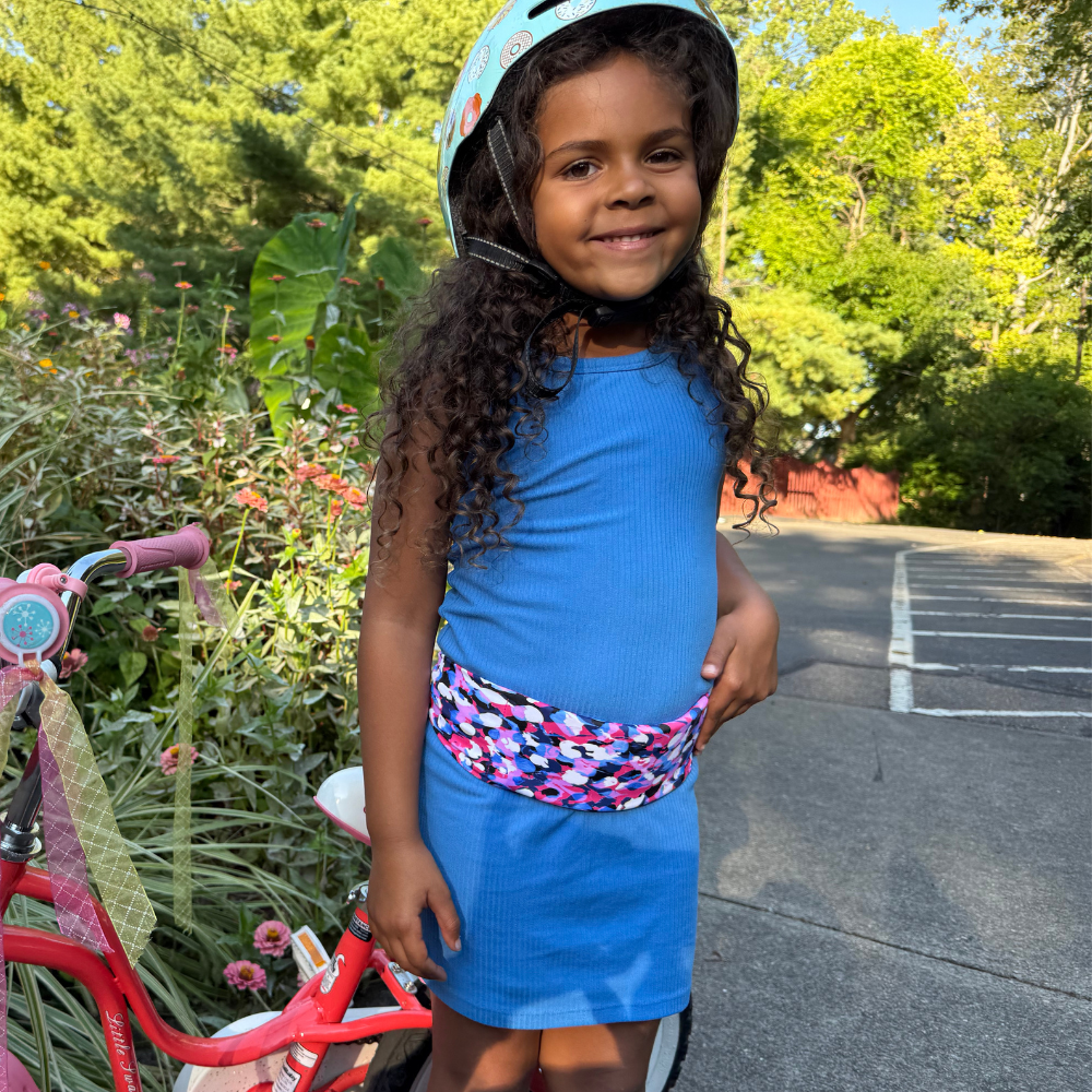 Young girl in a bike helmet and dress wearing a fanny pack in pink blue and white standing next to a pink bike outdoors.
